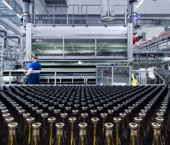 Produktionshalle mit einer Vielzahl an leeren Glasflaschen, die in Reihen angeordnet sind. Ein Mitarbeiter in blauer Arbeitskleidung überwacht den Prozess neben einer KHS-Maschine.