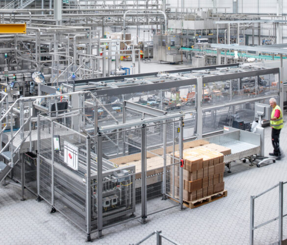 Production hall with packaging system and several conveyor belts. An employee in a safety vest works next to a stack of boxes.