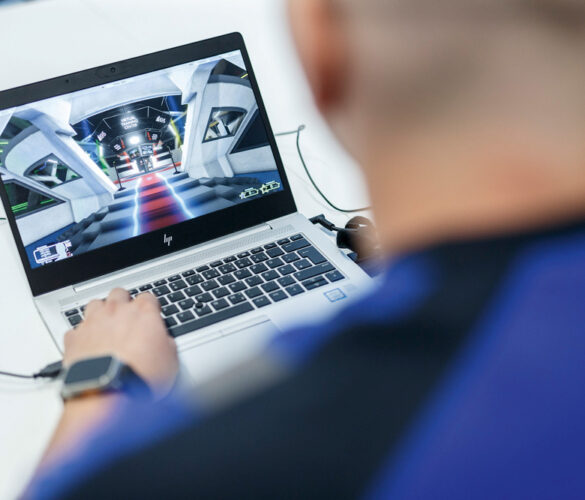 Person working on a laptop displaying a virtual environment of an industrial scene. The person is wearing a smartwatch on their wrist.